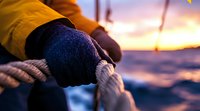 Gloved hands gripping nautical rope at sunset on boat, dramatic orange sky reflecting on ocean waves, maritime sailing atmosphere for adventure concepts.