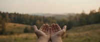 Open hands reaching toward golden sunset sky over peaceful countryside landscape during warm evening light.