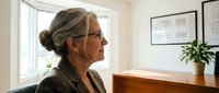 Mature woman with gray hair and glasses in profile, wearing olive blazer, sitting at wooden desk in bright office with window and plant.
