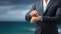 Businessman in gray suit checking time on wristwatch against blurred ocean background, conveying punctuality and professionalism.