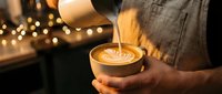 Barista pouring steamed milk creating latte art in coffee cup with warm bokeh lights background. Professional coffee making and cafe culture concept.