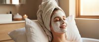 Young woman relaxing with white facial mask and towel on head in bright spa bathroom for skincare routine and self-care wellness treatment.