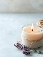 Glowing aromatic candle in glass jar decorated with twine, alongside fresh lavender sprigs on light textured background, creating peaceful spa atmosphere.