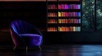 Luxurious velvet armchair in deep blue against dark interior with rainbow-colored book collection on shelves, moody atmospheric lighting creates cozy reading corner.