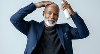 Mature African American male professional in navy blazer and gray turtleneck, hands raised to head in confident pose against white background for business and corporate use.