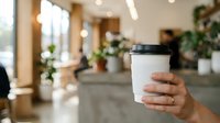 Hand holding white takeaway coffee cup in modern office lobby with plants and blurred coworkers in background, suitable for corporate, business, and workplace beverage concepts.