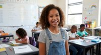 Happy young African American girl student smiling in elementary school classroom with diverse classmates studying at desks in bright educational environment.