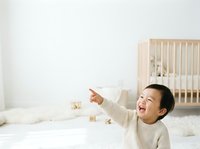 Young child with dark hair and East Asian ethnicity, wearing a cream-colored knit sweater, laughing and pointing upward in a bright, minimalist nursery with wooden crib and white bedding. Use for parenting, childcare, and family lifestyle content.