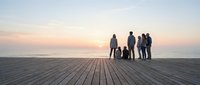 Group of diverse people standing together on wooden pier at sunset, silhouetted against ocean horizon. Team unity and connection concept.