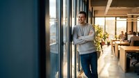 Confident young Hispanic businessman with crossed arms standing in modern office corridor with glass walls and natural lighting for corporate success concepts.