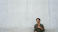 Young man with dark hair and facial hair, wearing olive green casual shirt, sitting against concrete wall with contemplative expression, suitable for lifestyle, fashion, and editorial content.
