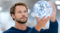 Young Caucasian man in navy sweater holding glowing digital sphere with network connections in modern office environment representing technology innovation.