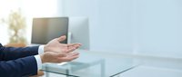 Professional businessman in dark suit extending hand for handshake in modern office with glass desk and computer, representing business partnership and deal.
