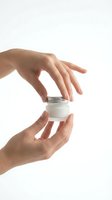 Female hands holding white cosmetic jar with metal lid against white background. Skincare product presentation for beauty, wellness, and cosmetics marketing.