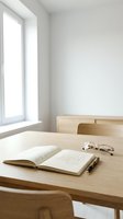 Open book with eyeglasses and pen on wooden desk in bright minimalist room with natural light from window, suitable for education and study concepts.