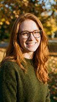 Happy young Caucasian woman with long red hair and glasses smiling outdoors in autumn park wearing green sweater during golden hour sunlight.