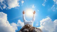 Person in white shirt raising hands towards bright sun in blue sky with white clouds, standing on mountain peak. Represents success, achievement, freedom and spirituality.