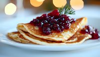 Homemade golden pancakes topped with sweet cranberry sauce and fresh dill garnish on white plate, festive bokeh lights in background for holiday breakfast.