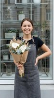Young woman with brown hair and fair complexion, wearing a dark gray apron and black t-shirt, smiling while holding a bouquet of white flowers and eucalyptus greenery wrapped in kraft paper in front of a modern plant shop storefront.
