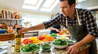 Middle-aged Caucasian male chef in plaid shirt and apron preparing fresh salad ingredients in bright modern kitchen with natural lighting from skylights.