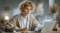 Young person with curly blonde hair and glasses working on laptop with digital data visualization effect, wearing beige cardigan in cozy home office setting.