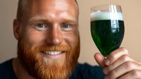 Smiling man with red beard holding glass of green beer or smoothie drink against neutral background, close up portrait showing genuine happiness.
