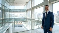 Confident male professional in dark business suit standing in modern high-rise office corridor with floor-to-ceiling windows overlooking city skyline, corporate environment.