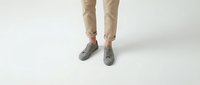 Cropped view of person wearing gray suede sneakers and beige rolled-up chinos, standing on white background, casual street style.