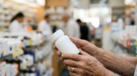 Close-up of mature male hands holding white medication bottle in pharmacy store with blurred shelves background for healthcare and pharmaceutical retail concepts.