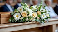 Elegant white and peach funeral flower arrangement on wooden casket during memorial service ceremony with mourners in background for sympathy condolences.