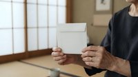 Senior man with weathered hands holding blank white envelope indoors, wearing dark clothing, suitable for mail, correspondence, voting, and communication concepts.