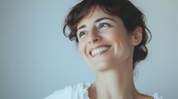 Young Caucasian woman with dark hair and bright genuine smile looking up with hopeful expression against light background, wearing white casual top. For wellness and positivity concepts.
