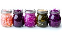 Colorful fermented vegetables in glass mason jars including pink radish, red cabbage, purple cabbage, cucumbers and onions arranged in row on white background.