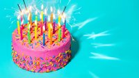 Festive pink birthday cake with colorful candles and sparklers on a bright teal background, covered in rainbow sprinkles.
