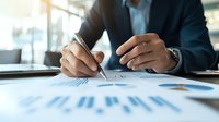 Businessman in dark suit analyzing financial charts and graphs, writing notes during data review and market research at modern office workspace.