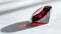 Sparkling red gemstone casting dramatic shadow on white surface, round brilliant cut ruby or garnet with deep crimson color and faceted clarity.