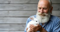 Senior man with long white beard and light skin tone, wearing blue shirt, holding white rat or mouse, isolated against wooden background for pet care, veterinary, and animal handling concepts.