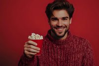 Young Caucasian man with beard wearing red knit sweater holding cup of popcorn, smiling against burgundy background. Casual winter lifestyle portrait.