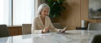 Mature woman with gray hair and light skin tone, wearing a beige blazer, reviewing documents at a marble conference table in a modern office setting.