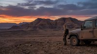 Adventurous woman standing beside rugged off-road vehicle in vast desert landscape during dramatic sunset with mountains and colorful sky.