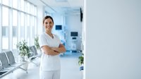 Confident female healthcare professional in white uniform standing with crossed arms in modern medical facility waiting area with natural lighting.