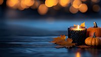 Glowing candle in black holder with small pumpkins and autumn maple leaves on dark wooden surface against blurred golden bokeh lights background.