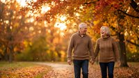 Happy senior couple walking hand in hand through autumn park with golden sunlight filtering through colorful fall foliage creating romantic atmosphere.