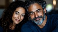 Mature multiethnic couple smiling at camera, middle-aged Hispanic man with salt and pepper beard and Mediterranean woman with natural makeup in casual setting.