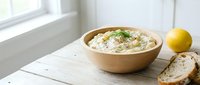 Creamy hummus dip in wooden bowl with fresh herbs, served with sliced bread and lemon on white table by window for food styling and culinary content.