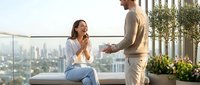 Young couple enjoying romantic moment on luxury rooftop terrace with city skyline view. Man proposing to surprised woman in casual attire on modern balcony.