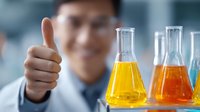 Young Asian male scientist showing thumbs up in laboratory with colorful chemical solutions in glass flasks, focus on yellow and orange liquids for scientific research.