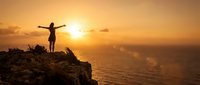 Woman silhouette with raised arms standing on rocky cliff edge during golden sunset over ocean waves, freedom and achievement concept for motivation.