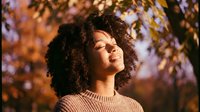 Young African American woman with curly hair enjoying peaceful moment outdoors in autumn sunlight with eyes closed and serene expression among fall foliage.