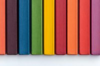 Colorful row of book spines arranged in rainbow order showing vibrant hues of red, blue, green, yellow, orange and pink against white background, creating minimalist pattern.